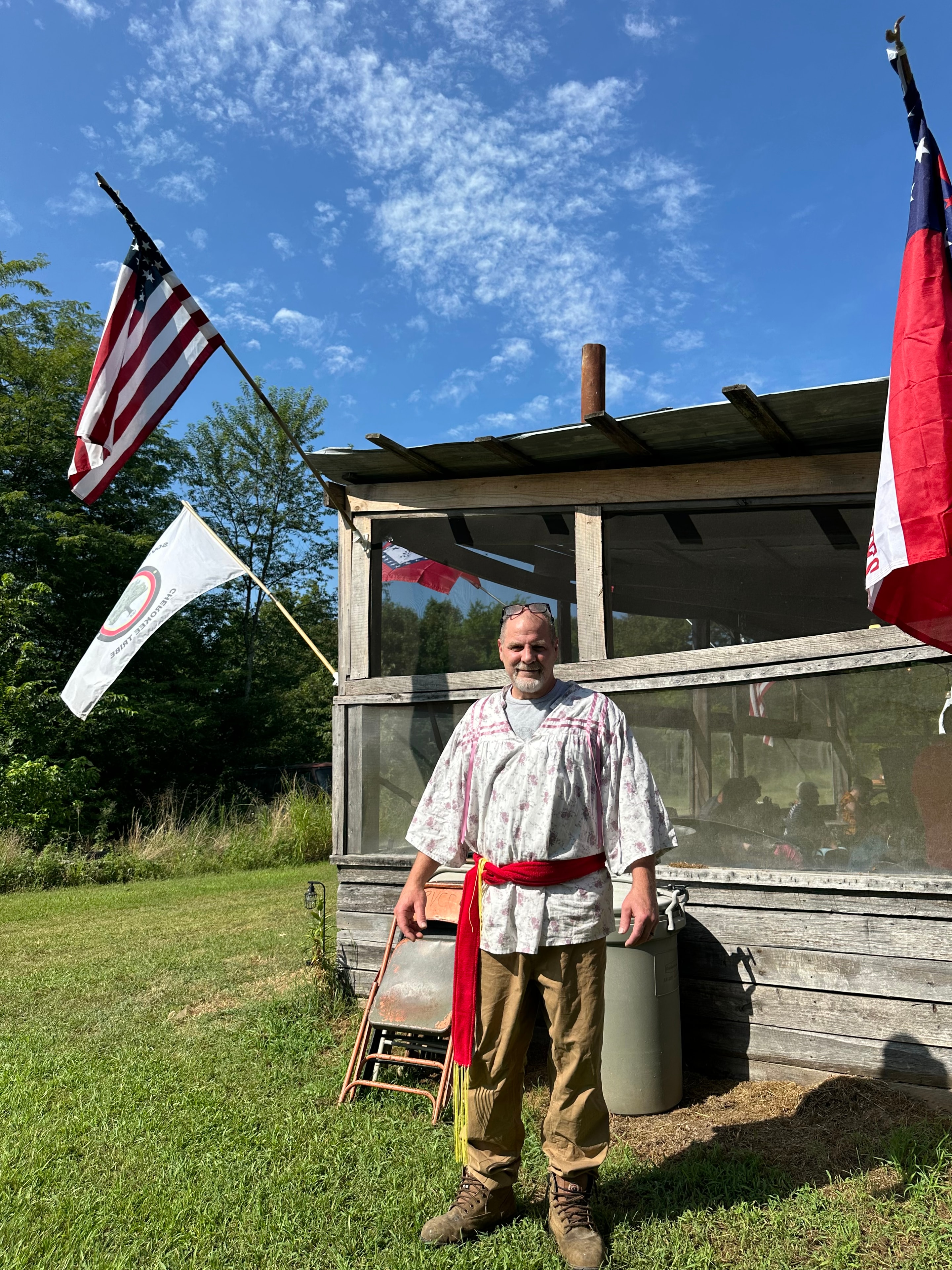 Tribal member in ribbon shirt standing by the ceremonial grounds with the American and tribal flags at Green Tree Ceremony July 11, 2026