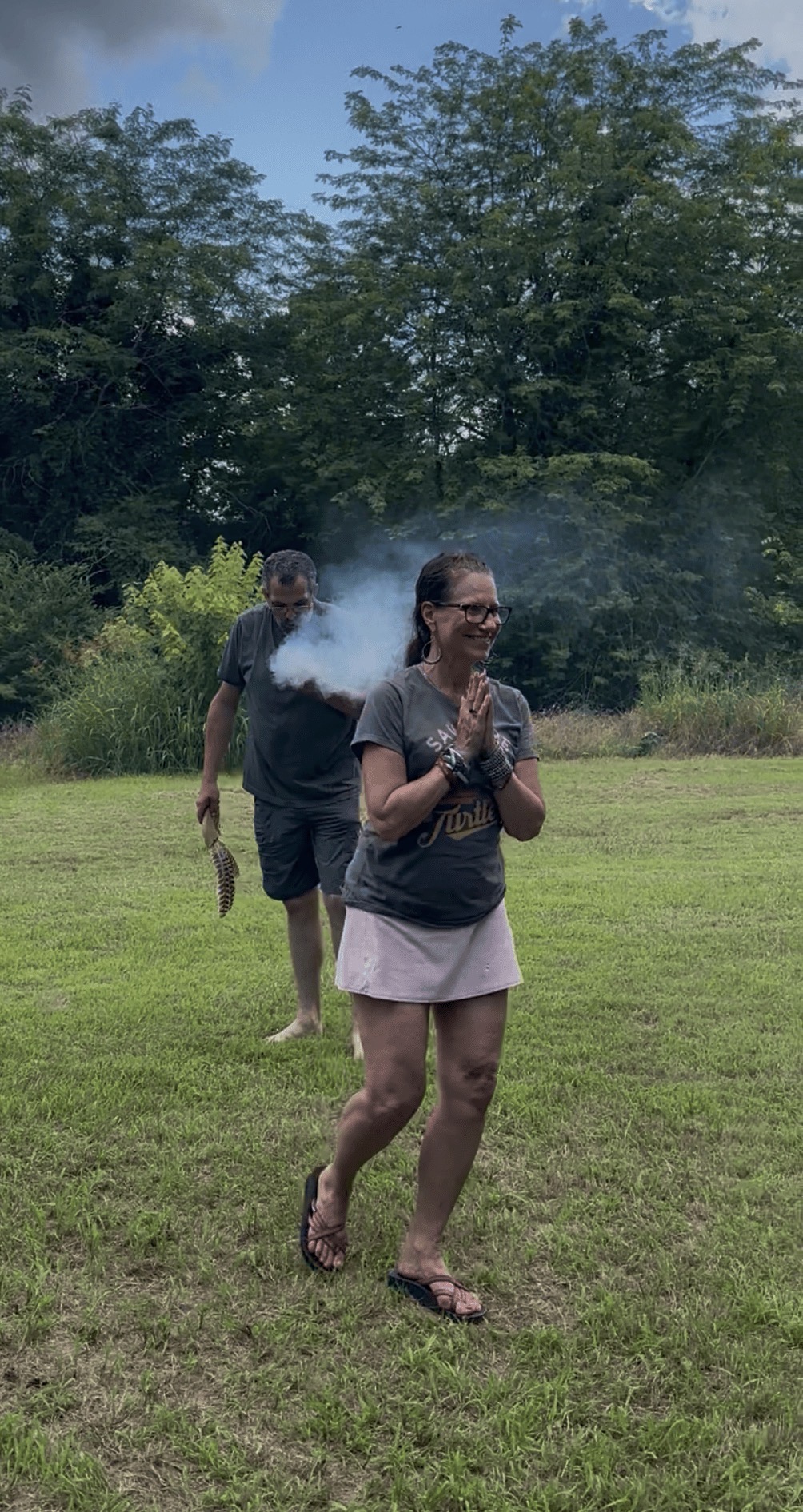 Tribal member smiling with hands in prayer as sacred smoke rises behind her at Green Corn Ceremony 2026